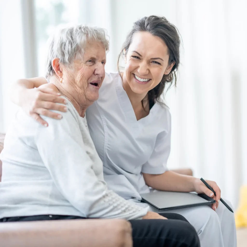 Alltagsbegleiterin sitzt lachend neben einer Seniorin auf dem Sofa