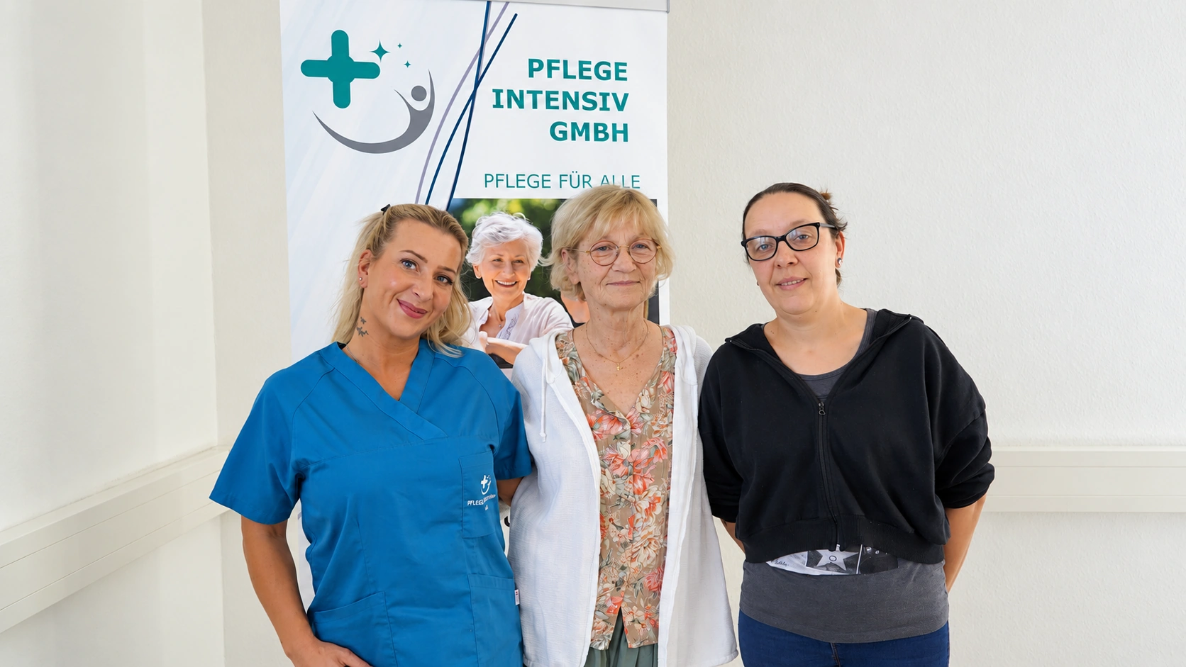Drei Mitarbeiterinnen der Pflege Intensiv GmbH stehen lächelnd vor dem Firmenaufsteller