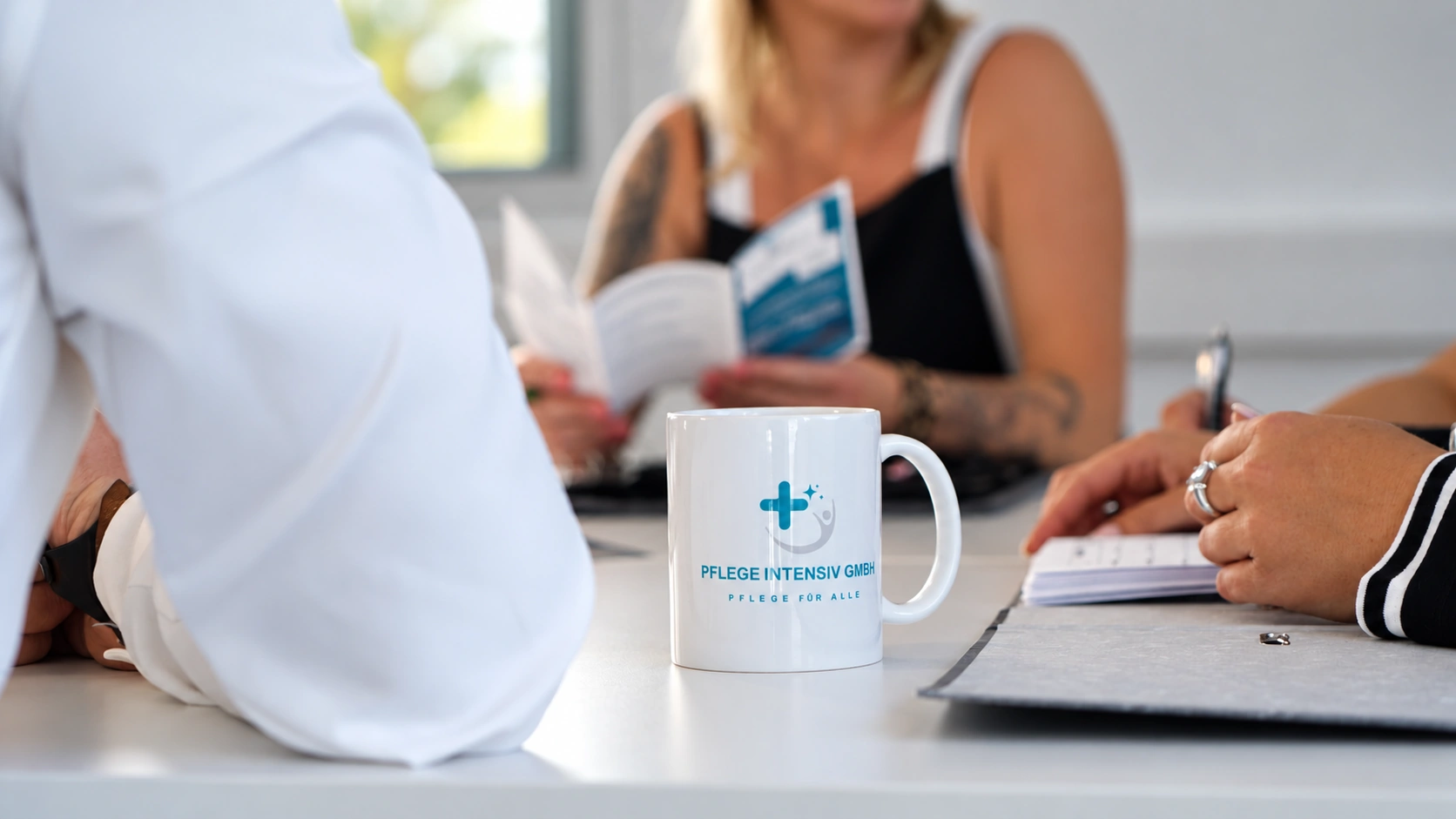 Tasse mit dem Logo der Pflege Intensiv GmbH auf dem Besprechungstisch, daneben Unterlagen und Notizen
