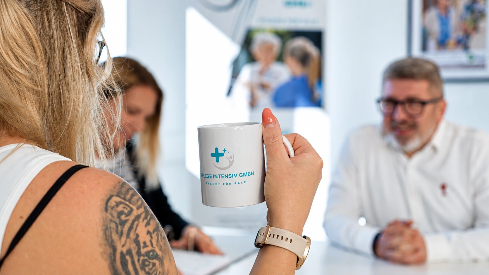 Mitarbeiterin mit Tasse der Pflege Intensiv GmbH in der Teambesprechung, im Hintergrund Kolleginnen und Kollegen