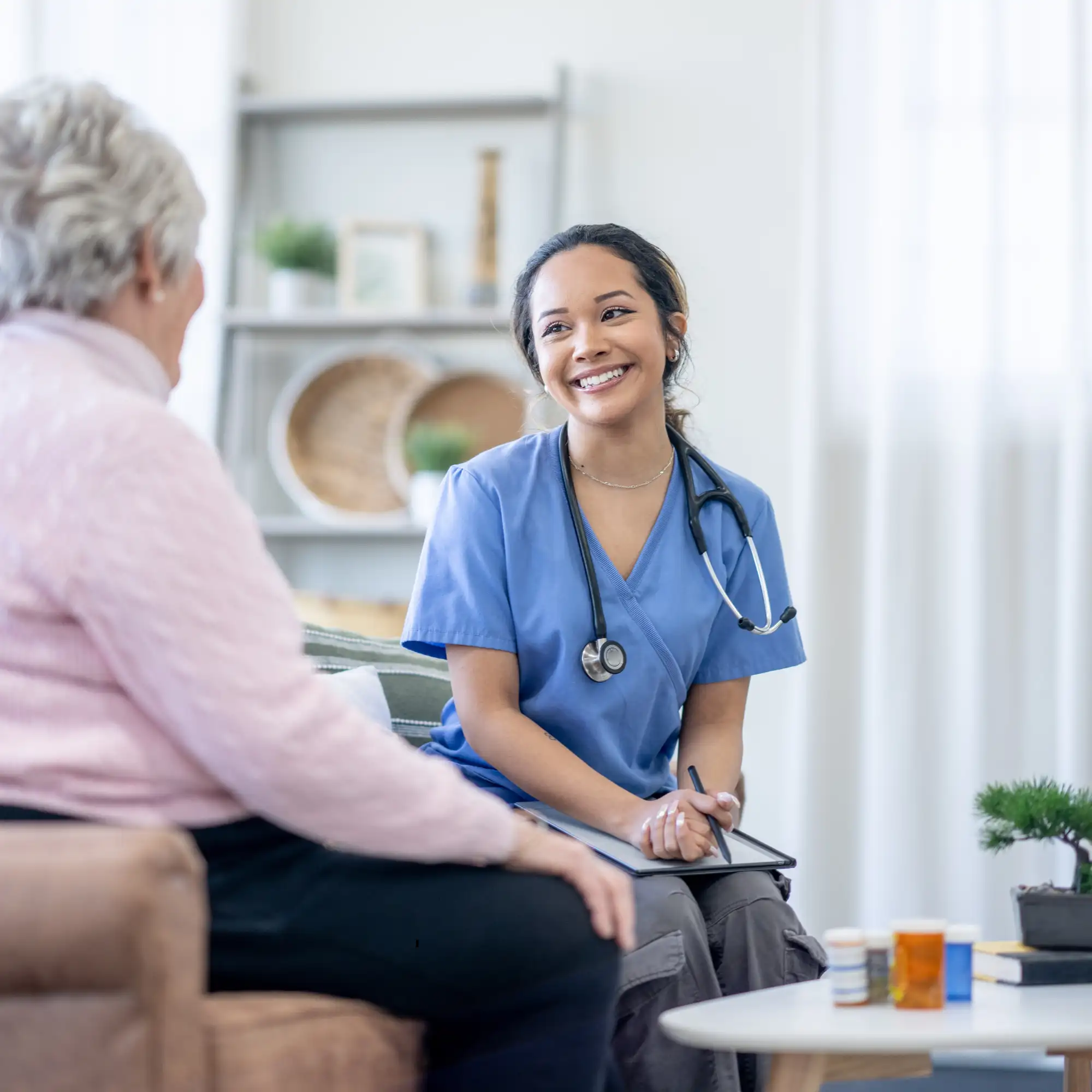 Junge Pflegekraft im blauen Kittel spricht lächelnd mit älterer Patientin in häuslicher Umgebung