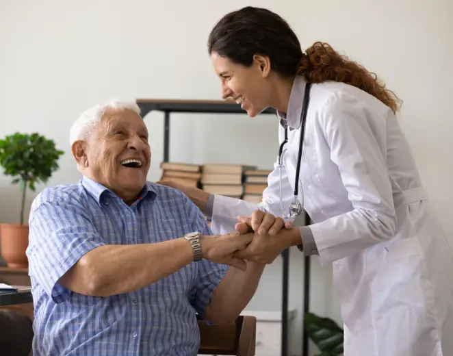 Pflegekraft in weißem Kittel hält lachend die Hände eines älteren Mannes – Zeit für persönliche Zuwendung