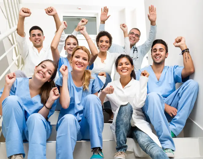 Pflege-Team sitzt lachend auf Treppe und reckt die Arme – Teamgeist und Zusammenhalt in der Pflege